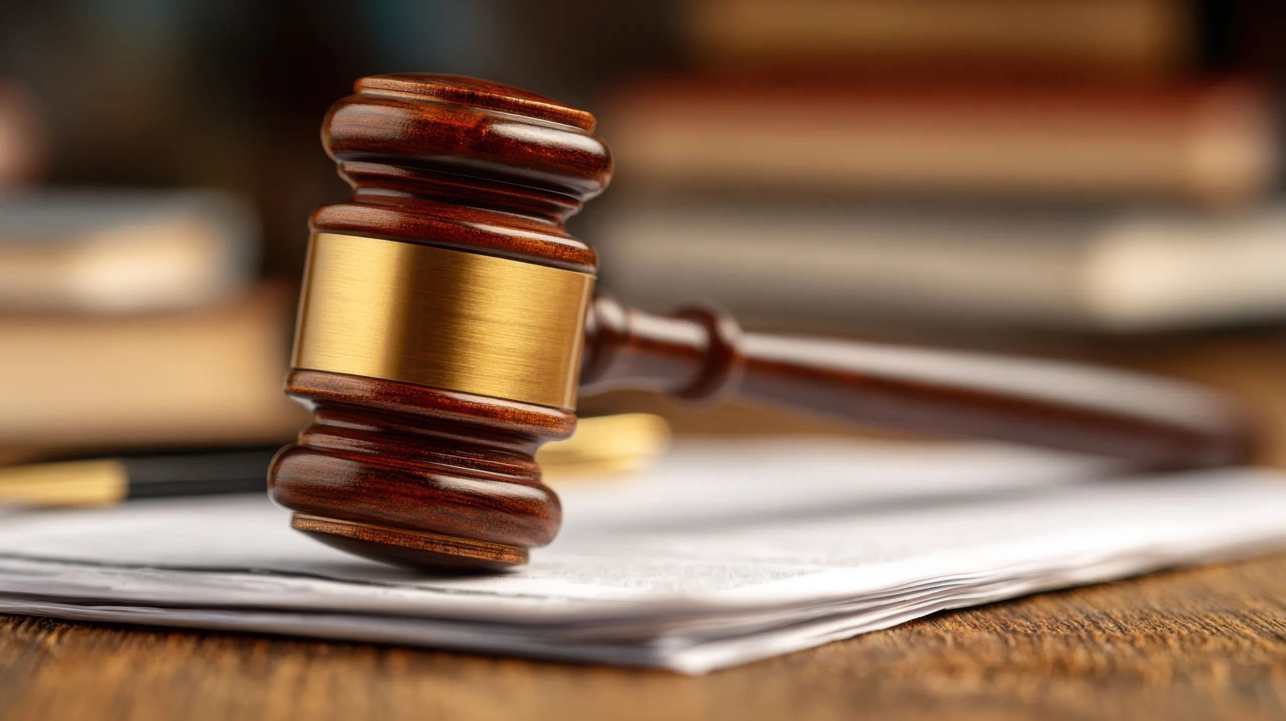 A wooden judge's gavel resting on a stack of papers on a desk with law books in the background